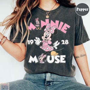 May include: Dark gray t-shirt featuring a Minnie Mouse graphic. The design includes the word "MINNIE" in pink, the year "1928", and the word "MOUSE" in white. Minnie Mouse is depicted in a pink polka dot dress.
