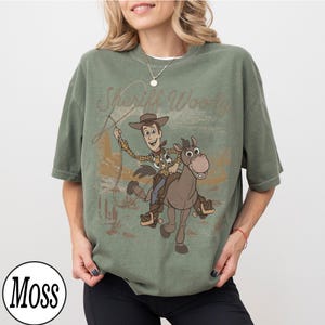 May include: An olive green t-shirt featuring a graphic of Sheriff Woody riding a horse with the text "Sheriff Woody". The design is in a vintage style. The shirt has short sleeves and a crew neck.