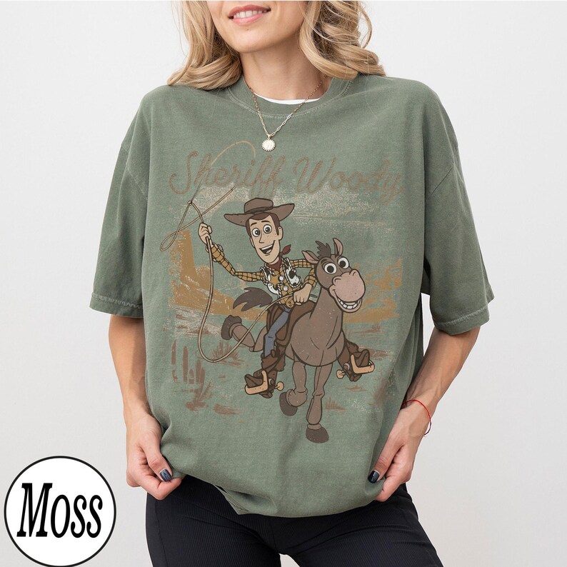May include: An olive green t-shirt featuring a graphic of Sheriff Woody riding a horse with the text "Sheriff Woody". The design is in a vintage style. The shirt has short sleeves and a crew neck.