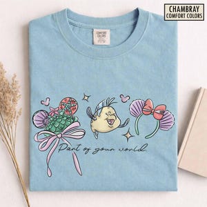 May include: Light blue t-shirt featuring a colorful graphic of a cartoon fish, a seashell-shaped headband with a pink bow, and a seashell-shaped design with a pink bow. The text "Part of your world" is also printed on the shirt.
