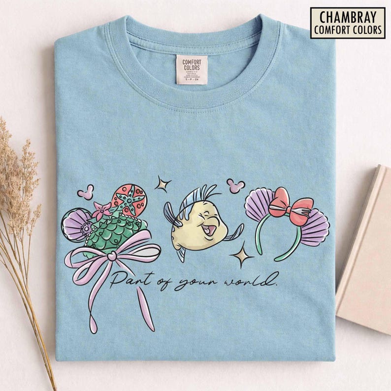 May include: Light blue t-shirt featuring a colorful graphic of a cartoon fish, a seashell-shaped headband with a pink bow, and a seashell-shaped design with a pink bow. The text "Part of your world" is also printed on the shirt.