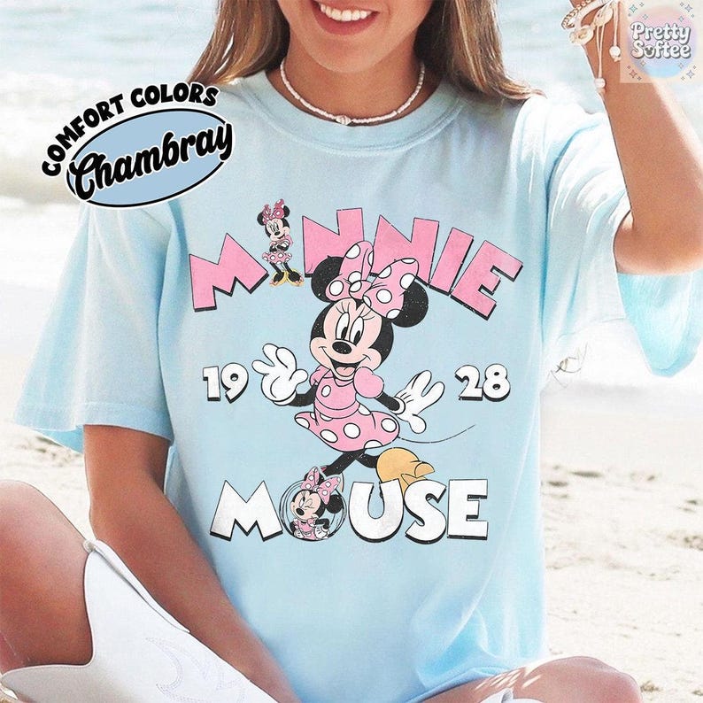 May include: Light blue t-shirt featuring Minnie Mouse in a pink polka-dot dress and bow. The shirt has the words "MINNIE MOUSE 1928" in pink and white. The shirt also has the words "COMFORT COLORS Chambray" in a circular design.