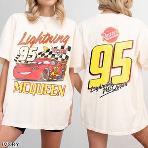May include: Ivory-colored t-shirt featuring a graphic of Lightning McQueen from the Cars movie. The front displays the text "Lightning McQueen 95" with a checkered flag and a red race car. The back shows a large yellow "95" and the text "Lightning McQueen".