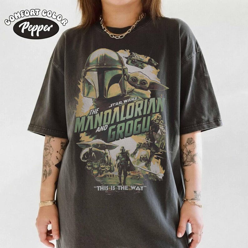 K&ouml;nnte beinhalten: Dunkelgraues T-Shirt mit einem Grafikdesign aus der Star Wars-Serie "The Mandalorian and Grogu". Das Design zeigt Bilder des Mandalorianers, Grogu und anderer Charaktere, mit dem Satz "THIS IS THE WAY" unten. Das Shirt hat das Comfort Colors-Logo.