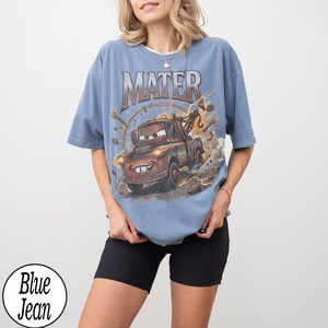 May include: A light blue t-shirt featuring a cartoon image of Mater from the movie Cars. The word "MATER" is printed across the top. The shirt has short sleeves and a round neckline. The words "Blue Jean" are printed on a white circle.