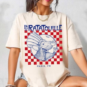 May include: Cream-colored t-shirt featuring a blue and red graphic design. The design includes the words "LA RATATOUILLE" in blue, a cartoon rat in a chef's hat, and the text "PARIS, FR". The background is a red and white checkered pattern.