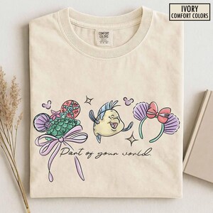 May include: Ivory-colored t-shirt with a whimsical design featuring cartoon elements. The design includes a seashell-shaped hat, a smiling fish, and a headband with a bow. The text "Part of your world" is also included.