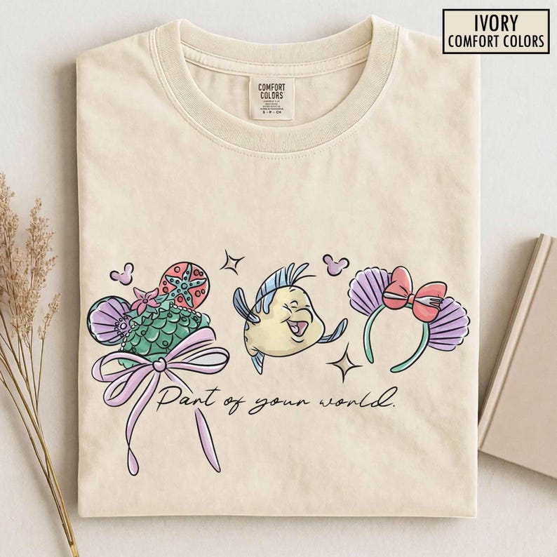 May include: Ivory-colored t-shirt with a whimsical design featuring cartoon elements. The design includes a seashell-shaped hat, a smiling fish, and a headband with a bow. The text "Part of your world" is also included.