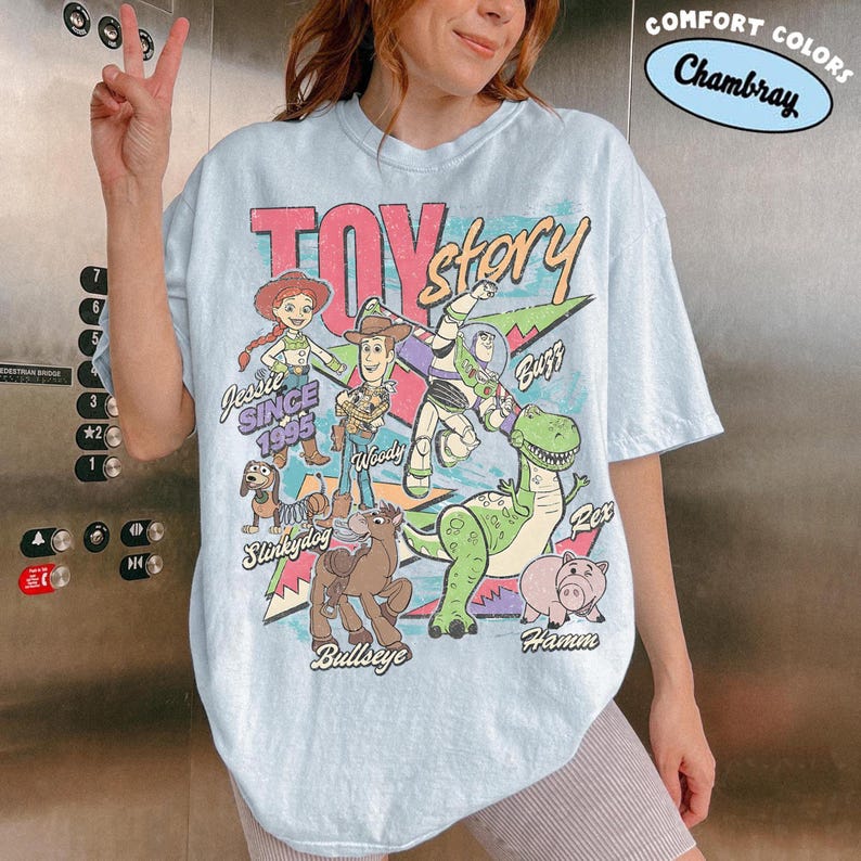 May include: Light blue t-shirt featuring a graphic of Toy Story characters, including Woody, Buzz, and Jessie. The shirt has the text "TOY STORY" and "SINCE 1995". The shirt is labeled "Comfort Colors Chambray".