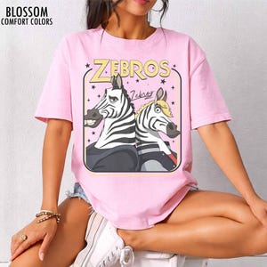 May include: Pink t-shirt with a cartoon graphic of two zebras. The graphic features the word "ZEBROS" in yellow, with a black outline, and the word "Zebras" in a cursive font. The shirt is made of a soft material and has short sleeves.