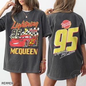 May include: Dark gray t-shirt featuring Lightning McQueen from the Cars movie. The front graphic shows McQueen with the number 95, a checkered flag, and the name McQueen. The back displays a large yellow 95 and the text "Lightning McQueen".