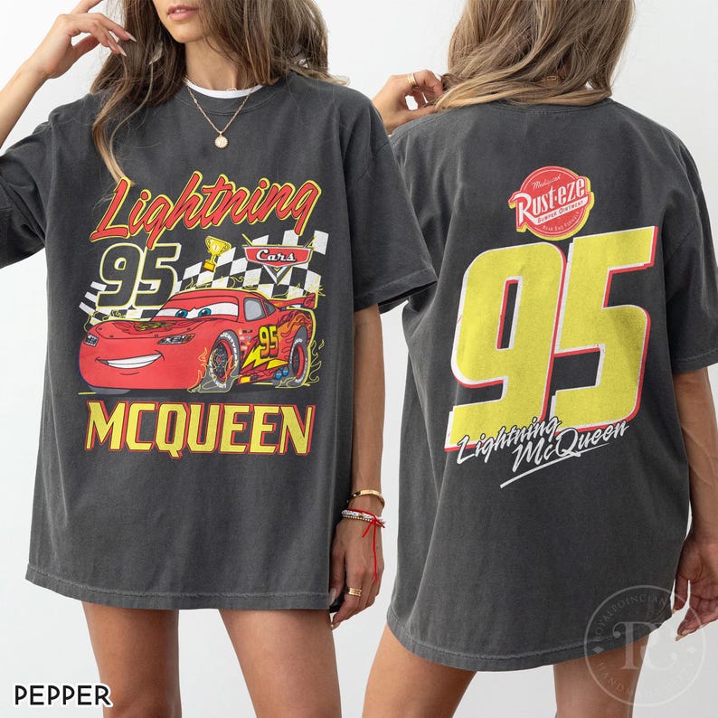 May include: Dark gray t-shirt featuring Lightning McQueen from the Cars movie. The front graphic shows McQueen with the number 95, a checkered flag, and the name McQueen. The back displays a large yellow 95 and the text "Lightning McQueen".