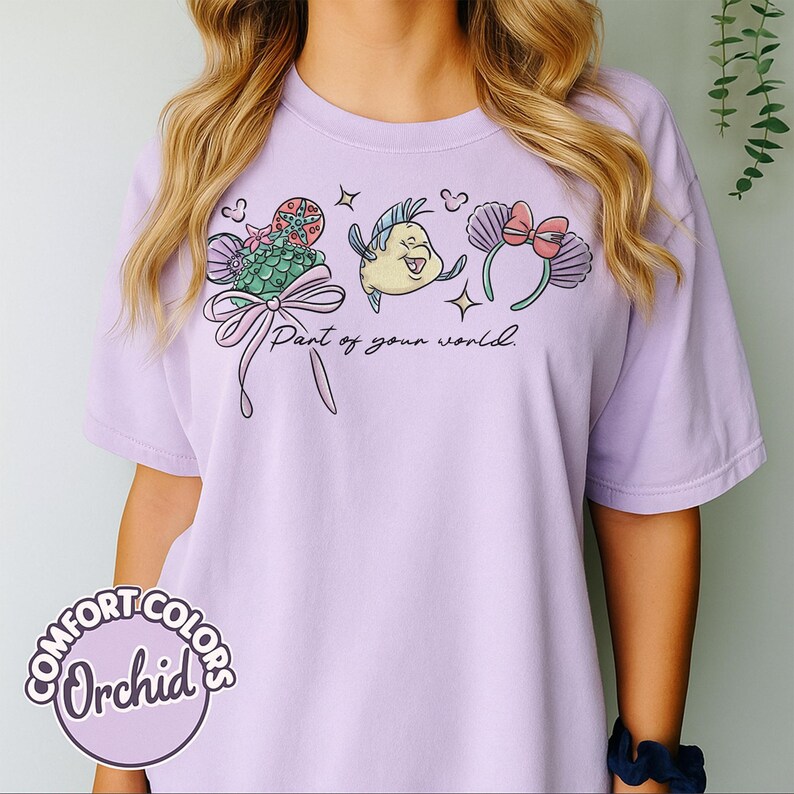 May include: Orchid-colored t-shirt featuring a whimsical illustration with the text "Part of your world." The design includes a mermaid tail, a fish, and a headband with a bow. The shirt has a relaxed fit and is made of a soft material.