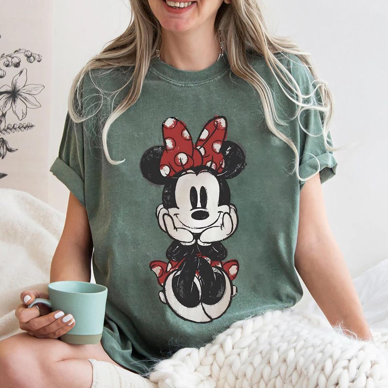 May include: Green t-shirt with a distressed Minnie Mouse graphic. The cartoon character is seated, wearing a red polka-dot bow and shoes. The shirt has rolled-up sleeves and a relaxed fit, suitable for casual wear.