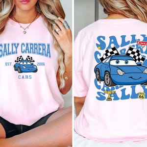以下が含まれることがあります： 水色のカートゥーンカーのデザインが施された、薄いピンク色のTシャツ。「SALLY CARRERA」と「CARS」の文字が前面にプリントされ、「EST. 2006」の文字も。背面には車、チェッカーフラッグ、そして「SALLY CARRERA」の文字が繰り返されています。