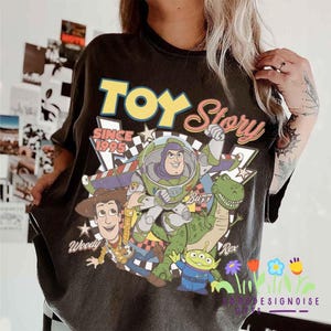 K&ouml;nnte beinhalten: Schwarzes T-Shirt mit einer farbenfrohen Grafik von Charakteren aus dem Animationsfilm "Toy Story". Das Design umfasst Woody, Buzz Lightyear, Rex und die kleinen gr&uuml;nen M&auml;nnchen, mit den Worten "Toy Story" und "Since 1995."