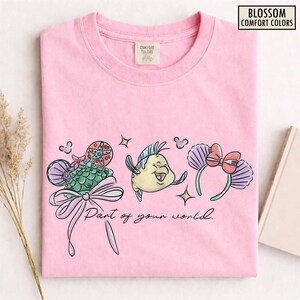 May include: A light pink t-shirt featuring a colorful graphic of a mermaid-themed design. The design includes a seashell, a fish, and a headband with a bow, along with the text "Part of your world."