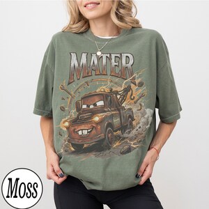 May include: Olive green t-shirt featuring a graphic of Mater from the movie Cars. The design includes the word "MATER" in a distressed font, lightning bolts, and tools. The shirt has a relaxed fit.