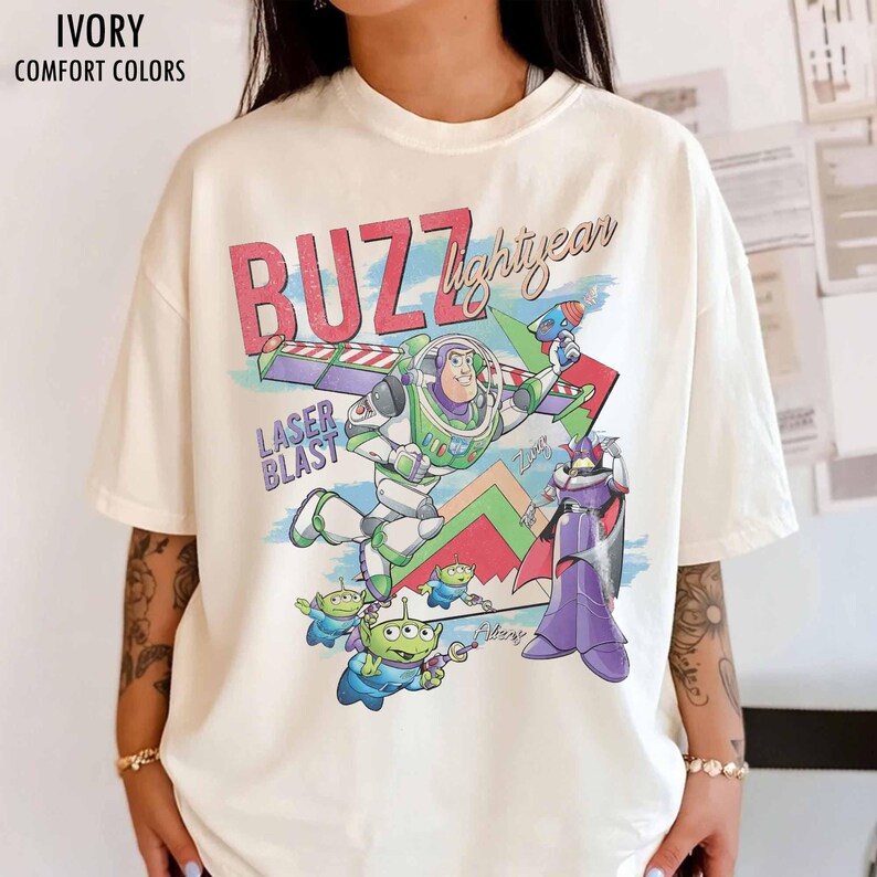 Puede incluir: Camiseta de color marfil con un gr&aacute;fico de Buzz Lightyear, extraterrestres y Zurg de la pel&iacute;cula Toy Story. La camiseta presenta las palabras "BUZZ lightyear" y "LASER BLAST" en un estilo retro. La camiseta est&aacute; etiquetada como "IVORY COMFORT COLORS".