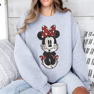 May include: A light grey sweatshirt with a distressed Minnie Mouse graphic. Minnie is shown with a red bow with white polka dots, a black body, and a smiling face. The sweatshirt has long sleeves and a crew neck.