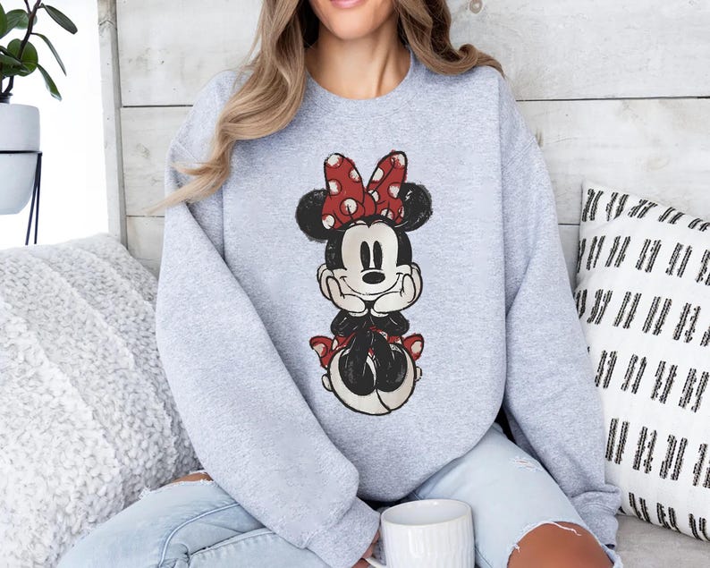 May include: A light grey sweatshirt with a distressed Minnie Mouse graphic. Minnie is shown with a red bow with white polka dots, a black body, and a smiling face. The sweatshirt has long sleeves and a crew neck.