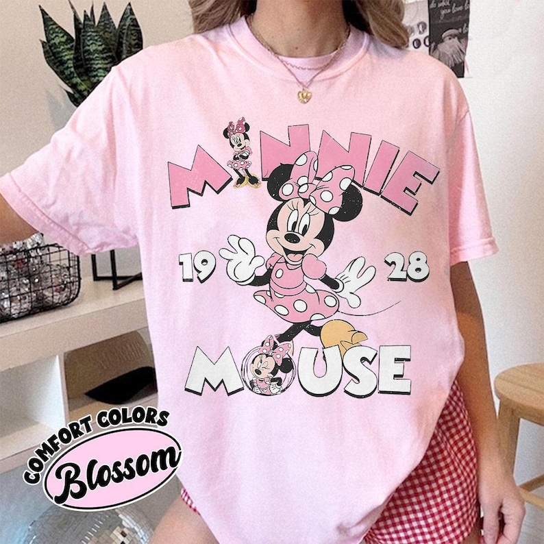 May include: A light pink t-shirt featuring a graphic of Minnie Mouse with the text "MINNIE MOUSE 1928". The shirt also has the text "COMFORT COLORS Blossom" in a circular design.