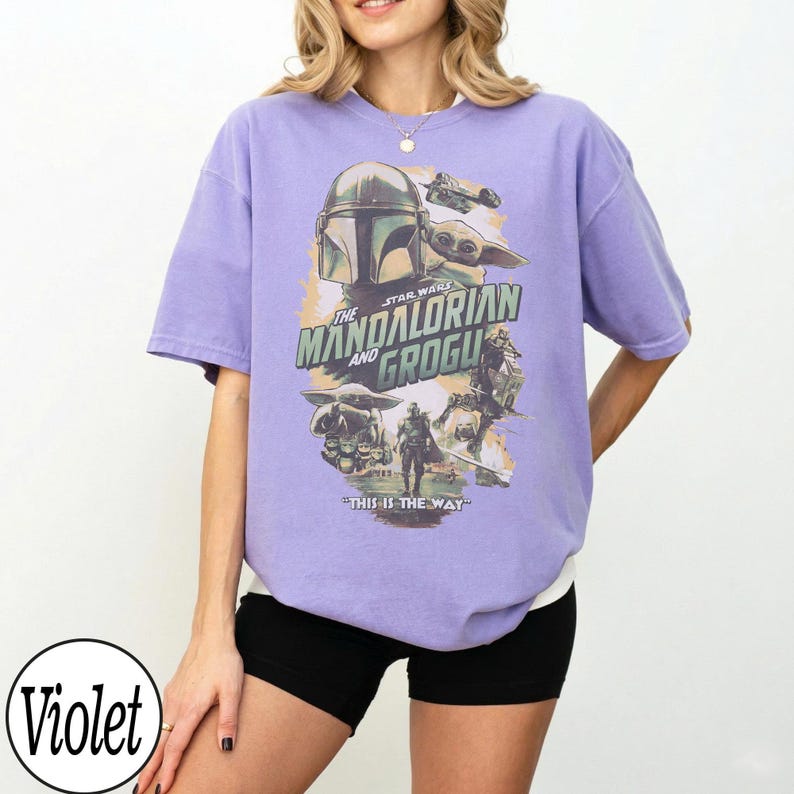 K&ouml;nnte beinhalten: Ein lila T-Shirt mit einem Grafikdesign aus der Serie "The Mandalorian". Das Design zeigt den Mandalorianer-Helm, Grogu und den Text "THE MANDALORIAN AND GROGU" und "THIS IS THE WAY". Das T-Shirt hat kurze &Auml;rmel und eine lockere Passform.