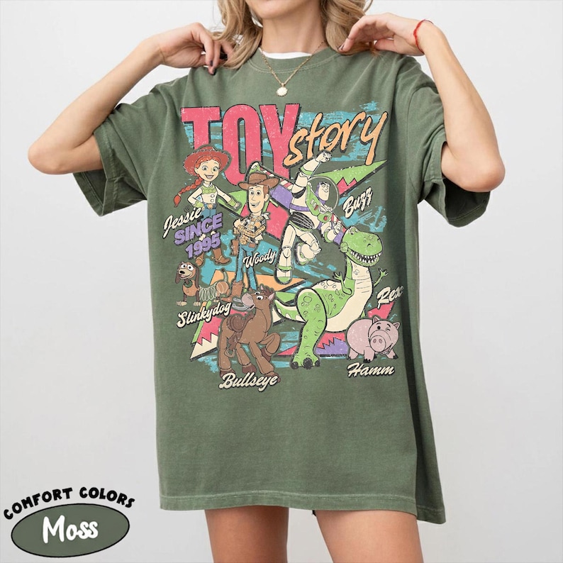 May include: A moss green t-shirt featuring a graphic of Toy Story characters, including Woody, Buzz Lightyear, and Jessie. The text "TOY STORY" is displayed above the characters, with "Jessie Since 1995" below. The shirt is labeled "Comfort Colors" and "Moss".