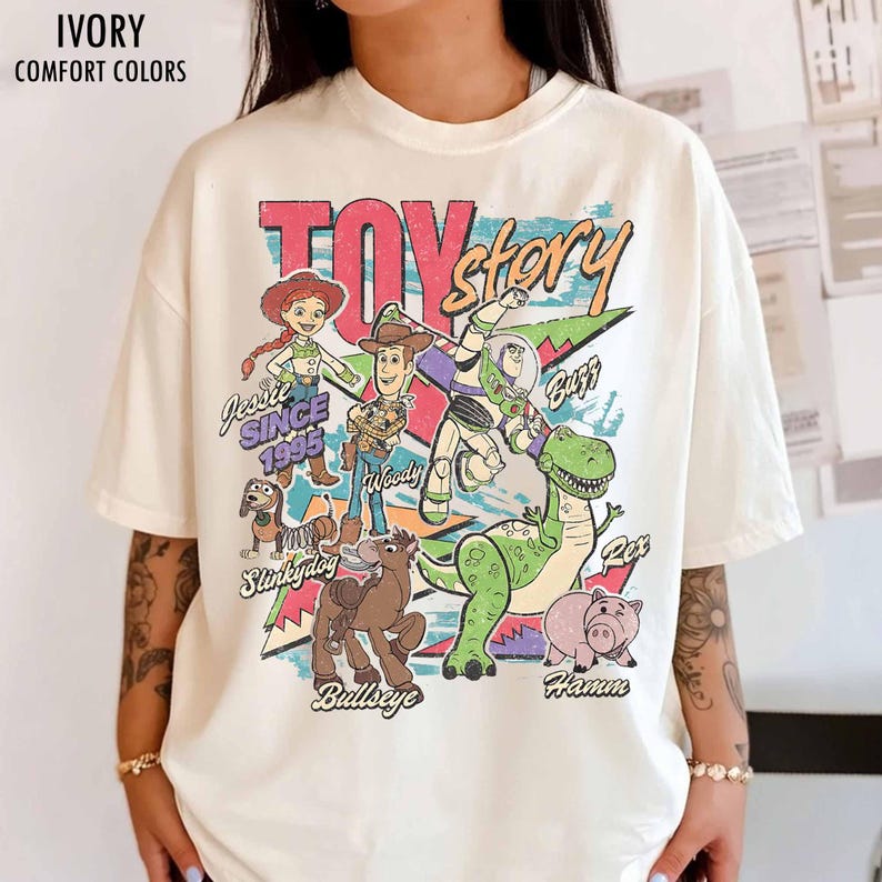 May include: Ivory-colored t-shirt featuring a colorful graphic of Toy Story characters, including Woody, Buzz Lightyear, and Rex. The text "TOY STORY" is prominently displayed, along with "Jessie Since 1995". The shirt has a relaxed fit.
