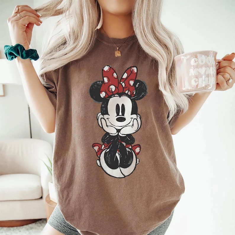 May include: A brown t-shirt with a Minnie Mouse graphic, featuring a red bow and black ears. The design is centered. The person is holding a pink mug with white text.