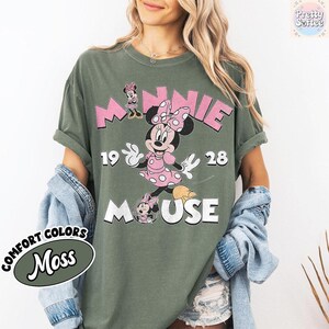 May include: Olive green t-shirt featuring a graphic of Minnie Mouse in a pink polka dot dress and bow. The text "MINNIE 1928 MOUSE" is printed above and below the character in pink and white. The shirt is labeled "Comfort Colors Moss".