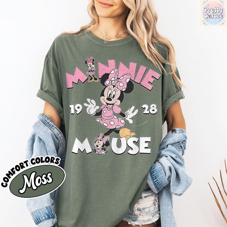 May include: Olive green t-shirt featuring a graphic of Minnie Mouse in a pink polka dot dress and bow. The text "MINNIE 1928 MOUSE" is printed above and below the character in pink and white. The shirt is labeled "Comfort Colors Moss".