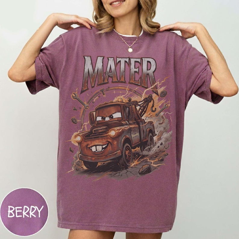 May include: A berry-coloured short-sleeve t-shirt featuring a graphic print of a rusty brown tow truck character with the text MATER arched above it. The design includes illustrated wrenches and mechanical parts surrounding the truck in a dusty setting.