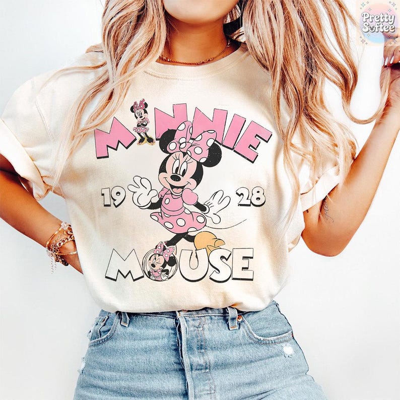 May include: Cream-colored t-shirt featuring a graphic of Minnie Mouse in a pink polka-dot dress and bow. The text "MINNIE 1928 MOUSE" is printed above and below the character in pink and black. The shirt has rolled-up sleeves.
