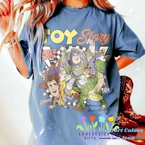 K&ouml;nnte beinhalten: Blaues T-Shirt mit einem Grafikdruck von Figuren aus dem Film "Toy Story". Das Design umfasst Buzz Lightyear, Woody, Rex und die kleinen gr&uuml;nen M&auml;nnchen, mit dem Text "Toy Story Since 1995."