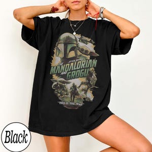 K&ouml;nnte beinhalten: Schwarzes T-Shirt mit einer Grafik von The Mandalorian und Grogu. Das Design zeigt den Helm des Mandalorianers, Grogu und den Text "The Mandalorian and Grogu" und "This is the Way".