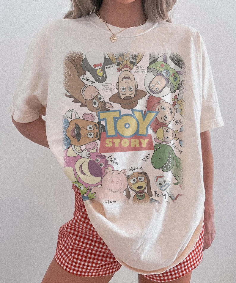 Może przedstawiać: Kremowy t-shirt z kr&oacute;tkim rękawem z dużym, kolorowym nadrukiem graficznym przedstawiającym postacie z Toy Story ułożone w okrąg wok&oacute;ł logo filmu. Wz&oacute;r zawiera postacie takie jak Woody, Buzz Astral, Jessie, Bystry, Rex, Hamm i inne.