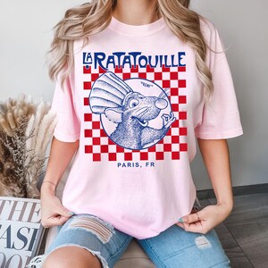 May include: A light pink t-shirt featuring a graphic from the movie "Ratatouille." The design includes the words "La Ratatouille" in blue, a cartoon rat, and the text "Paris, FR." The background has a red and white checkered pattern.