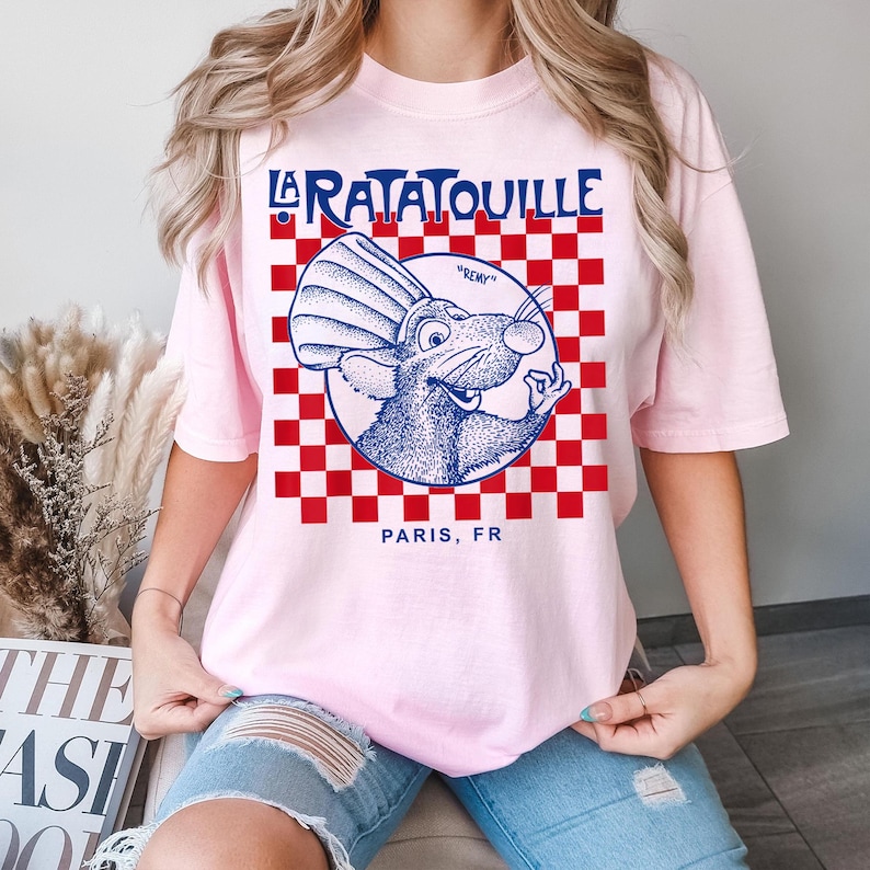 May include: A light pink t-shirt featuring a graphic from the movie "Ratatouille." The design includes the words "La Ratatouille" in blue, a cartoon rat, and the text "Paris, FR." The background has a red and white checkered pattern.