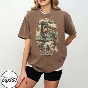 K&ouml;nnte beinhalten: Braunes T-Shirt mit einem Grafikdesign von "The Mandalorian" mit dem Text "The Mandalorian and Grogu" und "This is the Way". Das Design zeigt Bilder von Mandalorian, Grogu und anderen Charakteren aus der Star Wars-Serie.