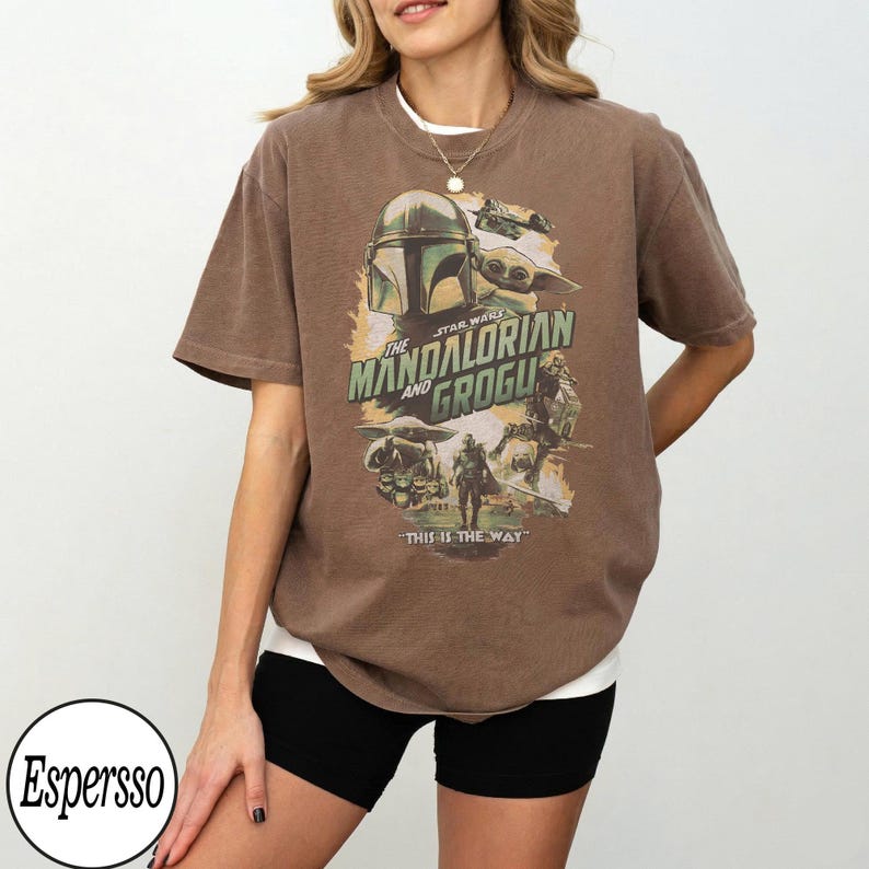 K&ouml;nnte beinhalten: Braunes T-Shirt mit einem Grafikdesign von "The Mandalorian" mit dem Text "The Mandalorian and Grogu" und "This is the Way". Das Design zeigt Bilder von Mandalorian, Grogu und anderen Charakteren aus der Star Wars-Serie.