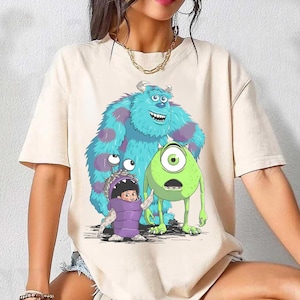 Puede incluir: Camiseta beige con un gráfico colorido de personajes de la película animada "Monstruos, S.A.". El diseño incluye a Sulley, Mike Wazowski y Boo con un disfraz de monstruo. La camiseta tiene mangas cortas y cuello redondo.