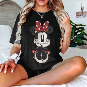 May include: Black t-shirt with a distressed Minnie Mouse design. The cartoon character has a red and white polka dot bow and matching details. The top is a classic crew neck style.