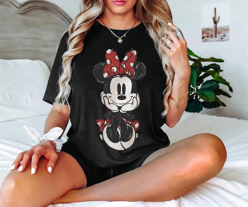 May include: Black t-shirt with a distressed Minnie Mouse design. The cartoon character has a red and white polka dot bow and matching details. The top is a classic crew neck style.