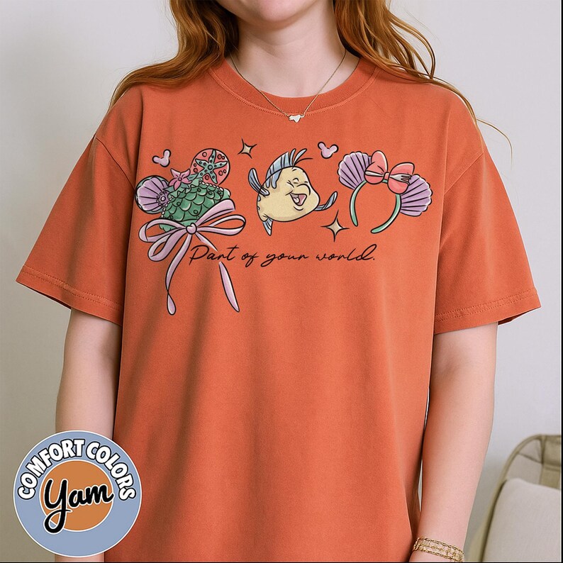 May include: Orange Comfort Colors t-shirt featuring a colorful graphic with the text "Part of your world." The graphic includes cartoon images of a fish, a flower, and a pair of headbands. The shirt has short sleeves and a round neckline.