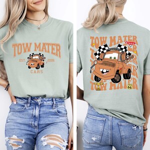 以下が含まれることがあります： 映画「カーズ」のキャラクター、メーターが描かれたライトグリーンのTシャツ。前面には「Tow Mater」の文字と、チェック柄の旗を持ったメーターのイラスト。背面には、メーターの大きな画像と「Tow Mater」の文字が繰り返されています。