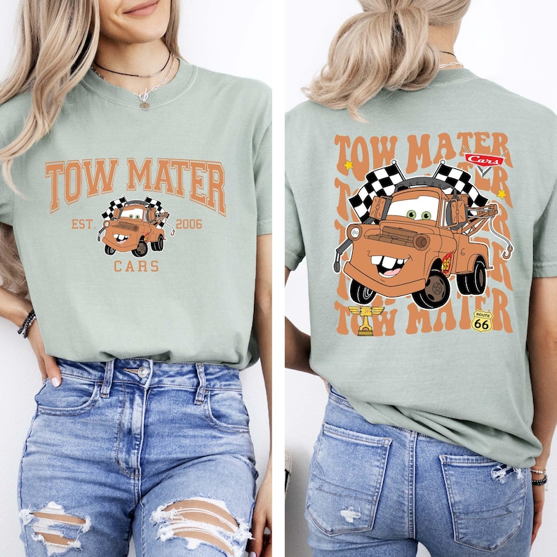 以下が含まれることがあります： 映画「カーズ」のキャラクター、メーターが描かれたライトグリーンのTシャツ。前面には「Tow Mater」の文字と、チェック柄の旗を持ったメーターのイラスト。背面には、メーターの大きな画像と「Tow Mater」の文字が繰り返されています。