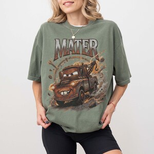 May include: An olive green short-sleeve t-shirt featuring a graphic print of a brown tow truck character with the text MATER above it. The design includes illustrations of mechanical tools like wrenches and gears surrounding the truck.