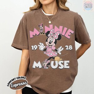 May include: Brown t-shirt featuring Minnie Mouse in a pink polka-dot dress and bow. The design includes the words "MINNIE MOUSE" and the numbers "1928". The shirt is labeled "Comfort Colors Espresso".