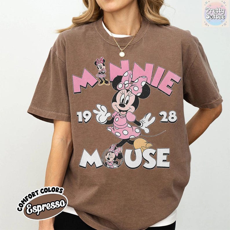 May include: Brown t-shirt featuring Minnie Mouse in a pink polka-dot dress and bow. The design includes the words "MINNIE MOUSE" and the numbers "1928". The shirt is labeled "Comfort Colors Espresso".